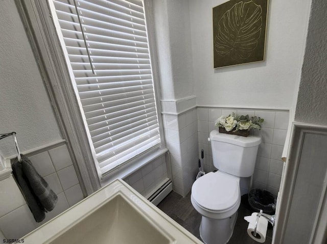 bathroom with baseboard heating, a textured wall, a wainscoted wall, toilet, and tile walls