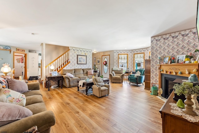 living area featuring wallpapered walls, light wood-type flooring, a glass covered fireplace, and stairway