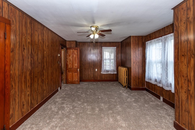 carpeted empty room featuring radiator heating unit, wooden walls, baseboards, and a ceiling fan