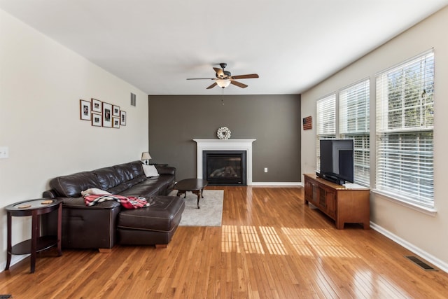 living area with a ceiling fan, a glass covered fireplace, visible vents, and light wood-style flooring