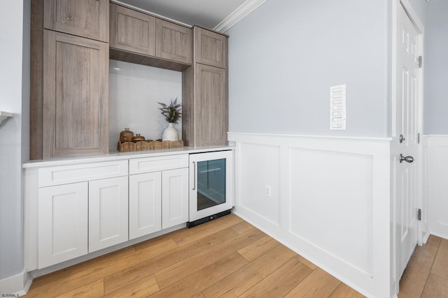 bar featuring crown molding, beverage cooler, light wood-type flooring, a decorative wall, and wainscoting