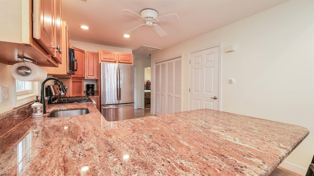 kitchen featuring freestanding refrigerator, a ceiling fan, a peninsula, light stone counters, and a sink