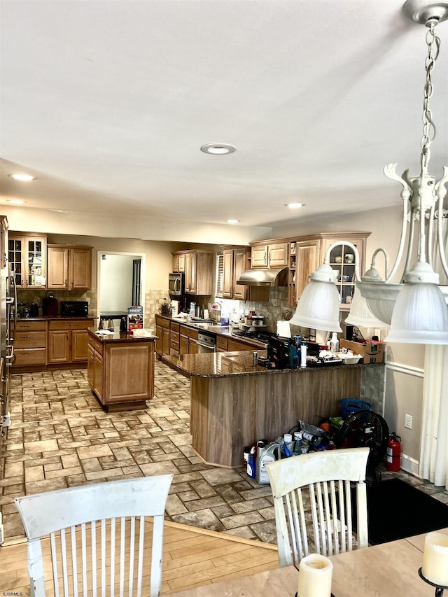 kitchen with glass insert cabinets, dishwashing machine, dark stone counters, under cabinet range hood, and backsplash
