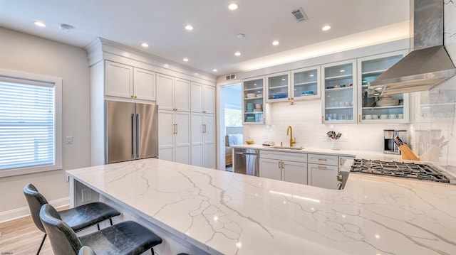 kitchen with glass insert cabinets, wall chimney range hood, tasteful backsplash, a sink, and appliances with stainless steel finishes
