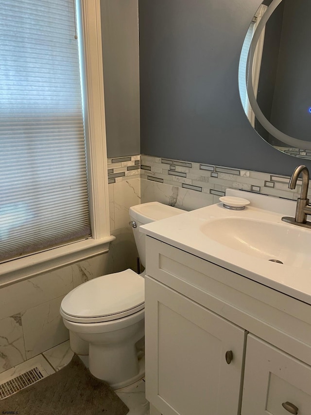 bathroom featuring tile walls, marble look tiles, toilet, vanity, and a wainscoted wall