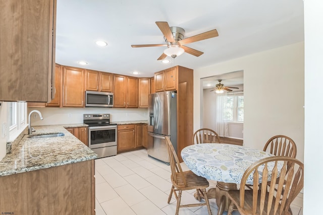 kitchen with appliances with stainless steel finishes, a sink, brown cabinets, light stone countertops, and a ceiling fan