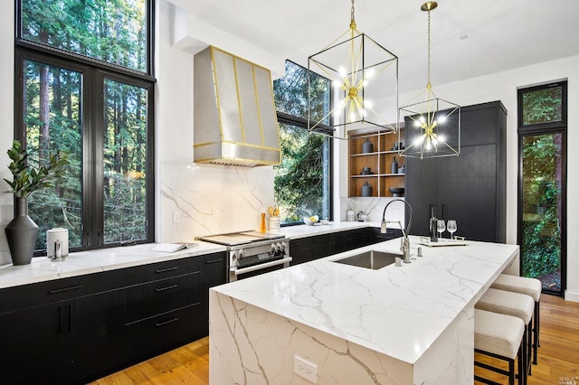 kitchen with wall chimney exhaust hood, sink, light wood-type flooring, a center island with sink, and tasteful backsplash