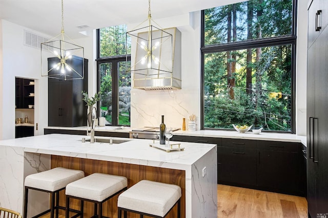 kitchen featuring decorative light fixtures, light wood-type flooring, a center island with sink, light stone counters, and wall chimney range hood