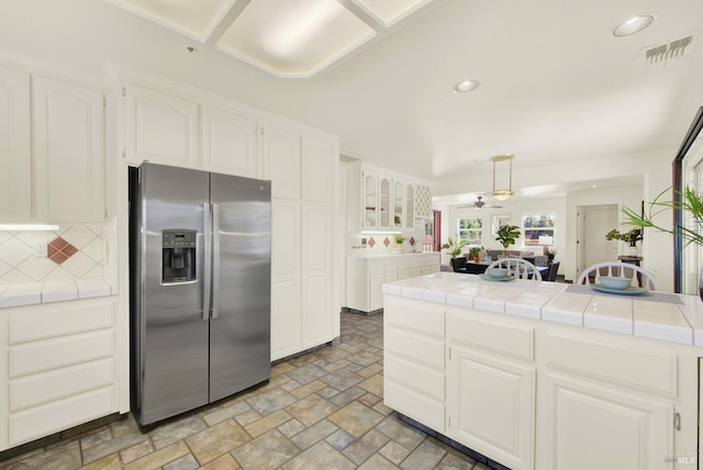 kitchen featuring backsplash, a ceiling fan, stone finish flooring, stainless steel fridge, and tile counters