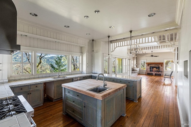 kitchen featuring a fireplace, ceiling fan with notable chandelier, dark wood-type flooring, a kitchen island with sink, and sink