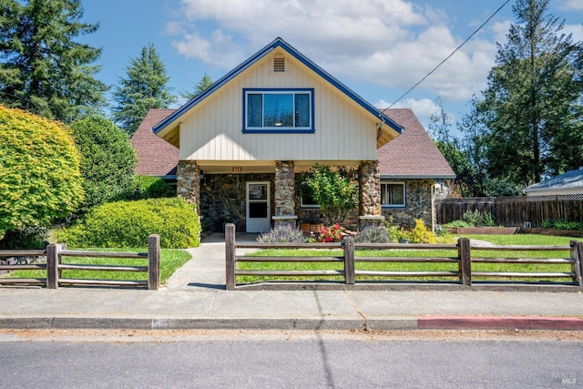 view of front of property featuring a fenced front yard, stone siding, covered porch, and roof with shingles