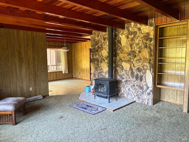 unfurnished living room with wooden walls, a wood stove, beamed ceiling, and carpet floors