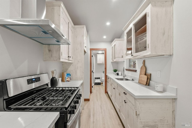 kitchen featuring stainless steel gas stove, wall chimney exhaust hood, light countertops, light wood-style floors, and a sink