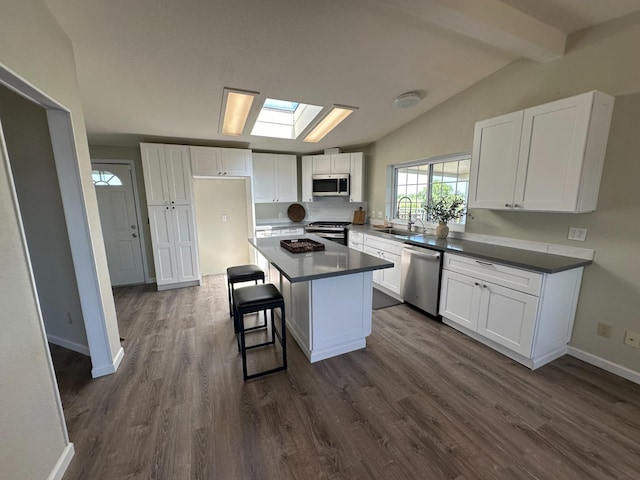 kitchen with vaulted ceiling with skylight, stainless steel appliances, a sink, white cabinets, and dark countertops