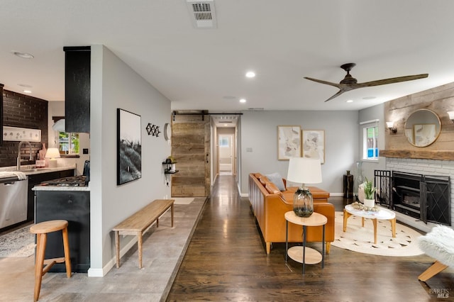 living room with dark wood-style flooring, visible vents, a barn door, a ceiling fan, and a brick fireplace