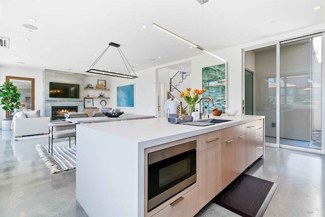 kitchen with light brown cabinets, decorative light fixtures, a large fireplace, a center island with sink, and stainless steel microwave