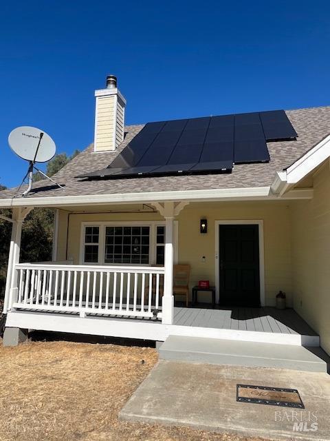 entrance to property featuring a porch, roof with shingles, a chimney, and solar panels