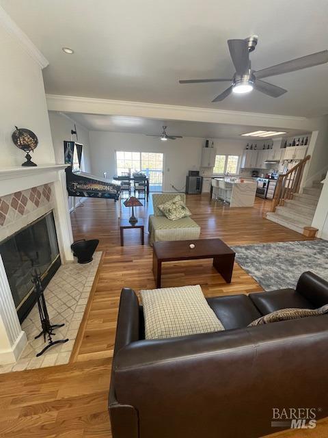 living area with ceiling fan, light wood-type flooring, a tiled fireplace, and crown molding