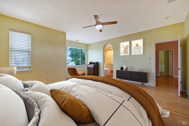 bedroom with light wood-type flooring, ensuite bathroom, baseboards, arched walkways, and ceiling fan