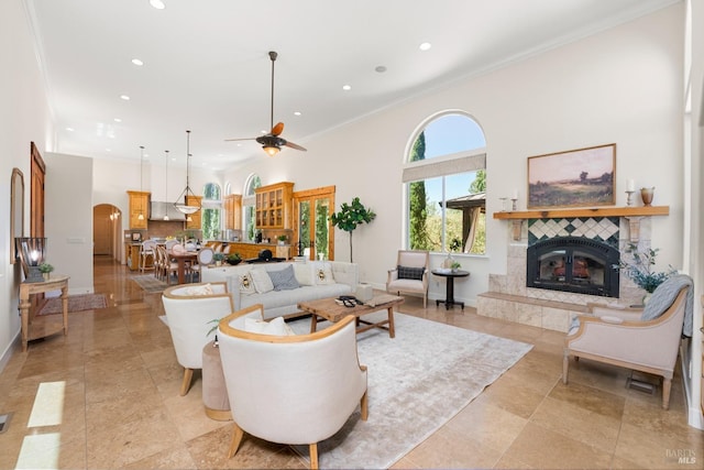 living room with arched walkways, recessed lighting, a towering ceiling, a tiled fireplace, and crown molding