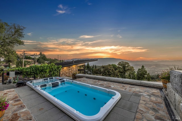 pool at dusk featuring a mountain view, an outdoor hot tub, and a patio area