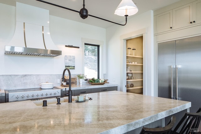 kitchen featuring stainless steel built in refrigerator, wall chimney exhaust hood, hanging light fixtures, and a kitchen bar