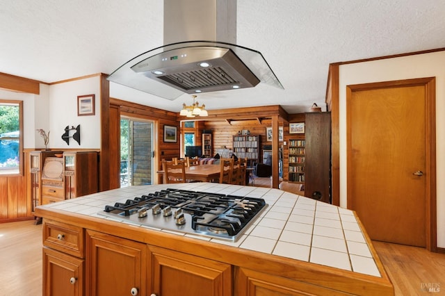 kitchen with tile counters, light hardwood / wood-style floors, wood walls, island range hood, and stainless steel gas stovetop