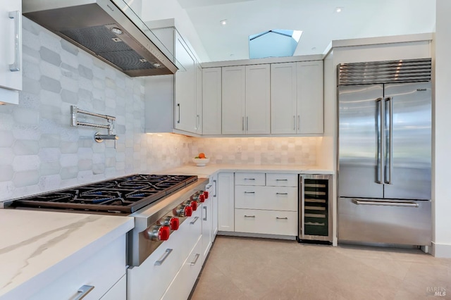 kitchen with appliances with stainless steel finishes, white cabinets, beverage cooler, exhaust hood, and light stone counters