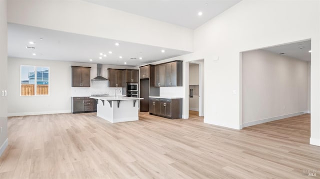 kitchen with appliances with stainless steel finishes, light hardwood / wood-style flooring, a kitchen breakfast bar, wall chimney exhaust hood, and a kitchen island with sink