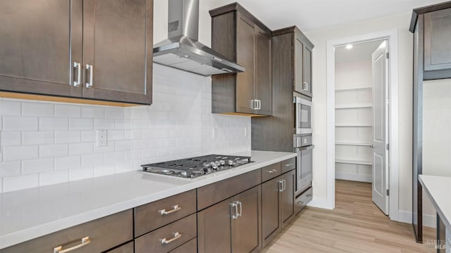 kitchen with stainless steel appliances, light hardwood / wood-style flooring, dark brown cabinets, tasteful backsplash, and wall chimney range hood