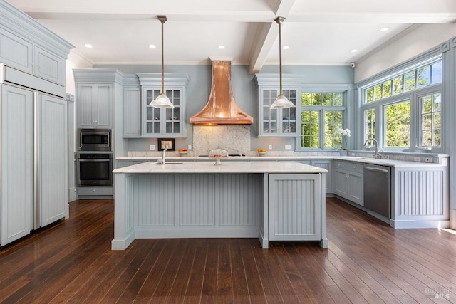 kitchen with dark wood-type flooring, hanging light fixtures, built in appliances, an island with sink, and custom exhaust hood