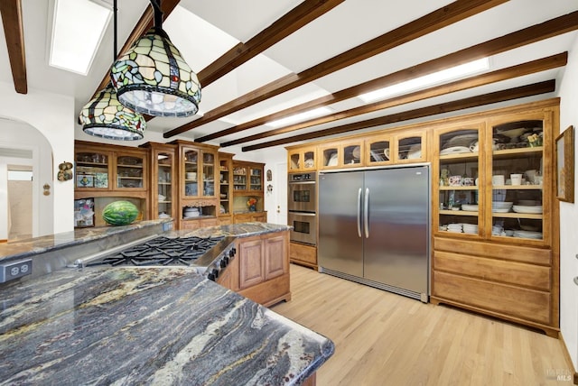 kitchen with appliances with stainless steel finishes, dark stone countertops, beam ceiling, and light hardwood / wood-style floors