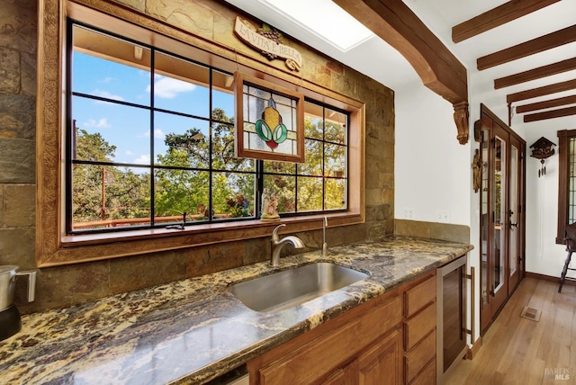 kitchen featuring sink, light wood-type flooring, beamed ceiling, dark stone counters, and beverage cooler