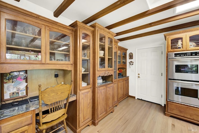 kitchen featuring double oven, built in desk, light hardwood / wood-style floors, dark stone counters, and beamed ceiling