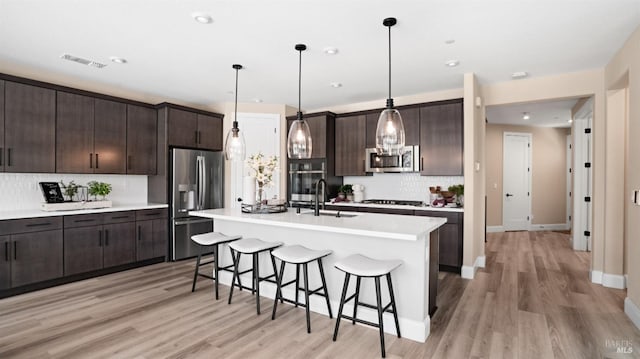 kitchen with dark brown cabinetry, sink, pendant lighting, stainless steel appliances, and a kitchen island with sink