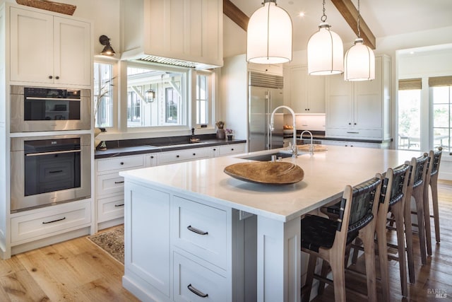 kitchen featuring stainless steel appliances, white cabinetry, decorative light fixtures, an island with sink, and light wood-type flooring