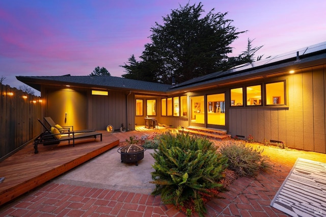 back house at dusk with a wooden deck, a patio area, solar panels, and an outdoor fire pit