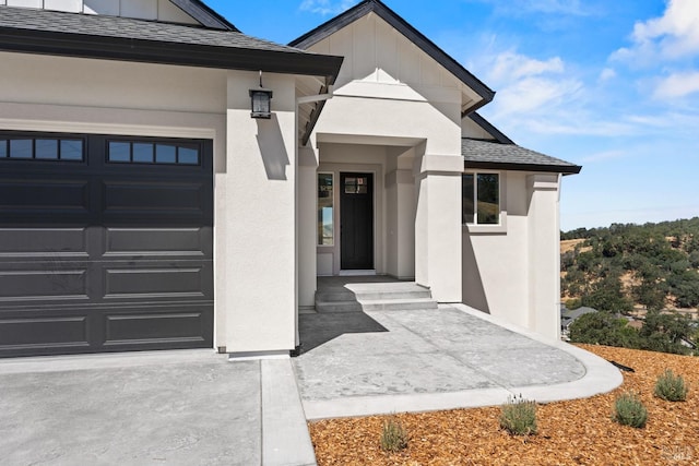 view of exterior entry with an attached garage, stucco siding, board and batten siding, and roof with shingles