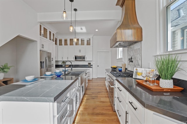 kitchen with appliances with stainless steel finishes, custom exhaust hood, a large island with sink, decorative light fixtures, and white cabinets