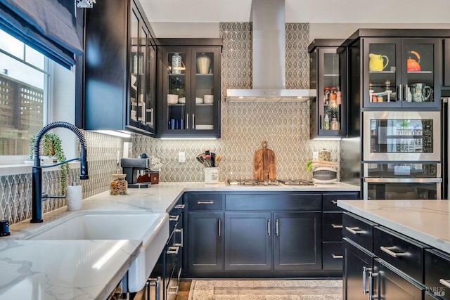 bar with light stone countertops, sink, stainless steel appliances, wall chimney range hood, and tasteful backsplash