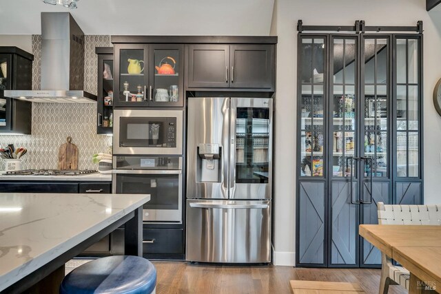 kitchen with hardwood / wood-style flooring, wall chimney exhaust hood, light stone counters, dark brown cabinetry, and stainless steel appliances