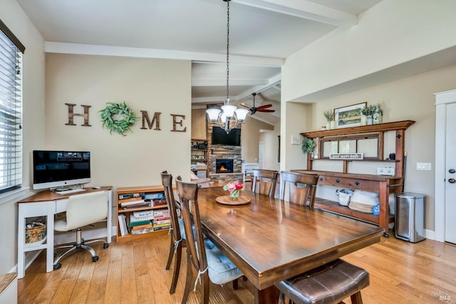 dining room featuring lofted ceiling with beams, a stone fireplace, light wood-type flooring, and ceiling fan with notable chandelier