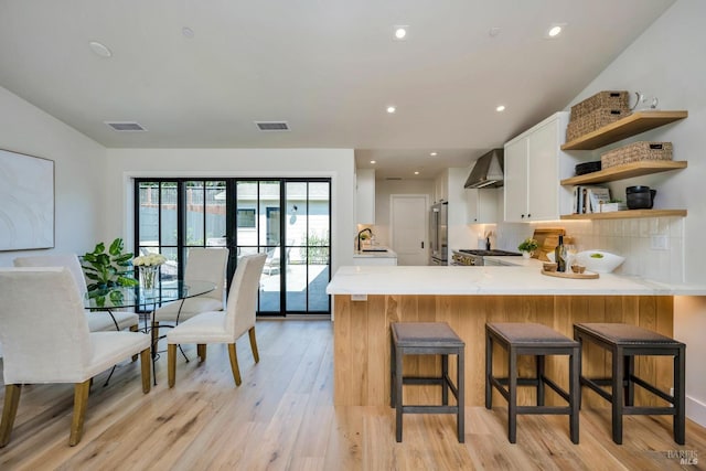 kitchen featuring a peninsula, light countertops, wall chimney range hood, white cabinetry, and open shelves