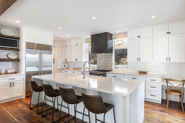 kitchen with range hood, backsplash, light countertops, stainless steel built in refrigerator, and a breakfast bar area