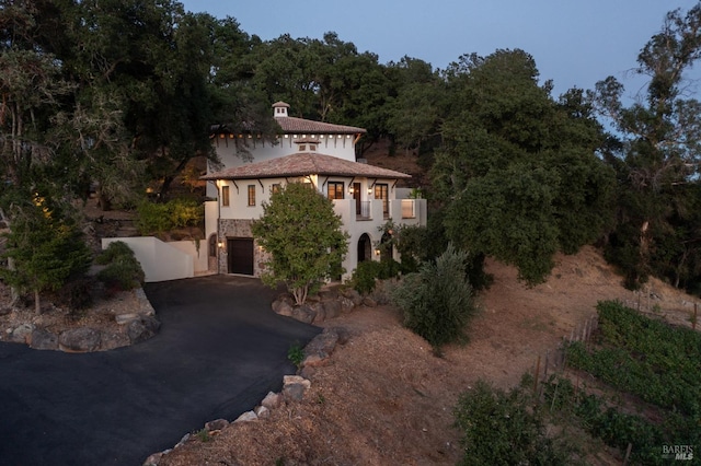 mediterranean / spanish house featuring aphalt driveway, stone siding, a view of trees, an attached garage, and stucco siding
