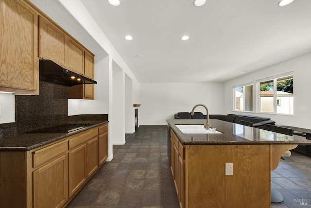 kitchen with dark stone countertops, sink, an island with sink, black electric cooktop, and a breakfast bar