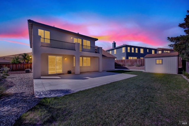 back house at dusk featuring a balcony, a patio area, a lawn, and a shed