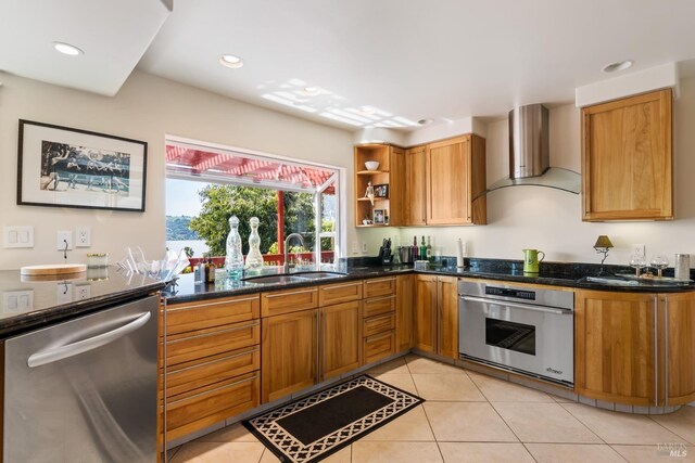 kitchen with wall chimney exhaust hood, light tile patterned floors, appliances with stainless steel finishes, sink, and dark stone counters