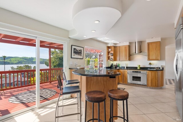 kitchen with wall chimney exhaust hood, light tile patterned floors, a center island, stainless steel appliances, and a water view