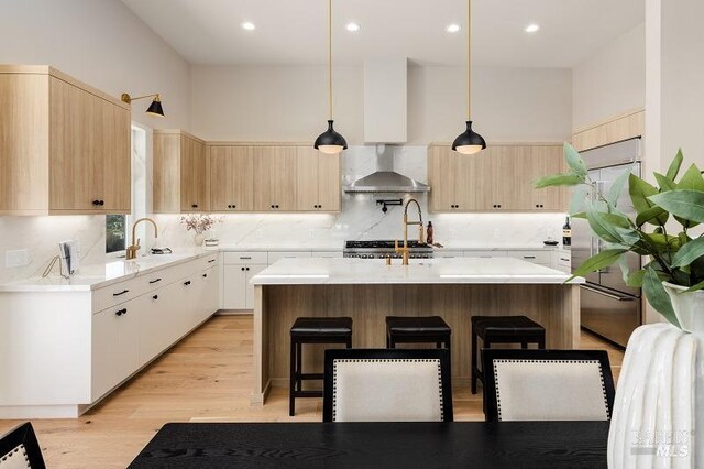 kitchen featuring a center island with sink, light hardwood / wood-style floors, built in refrigerator, hanging light fixtures, and wall chimney range hood